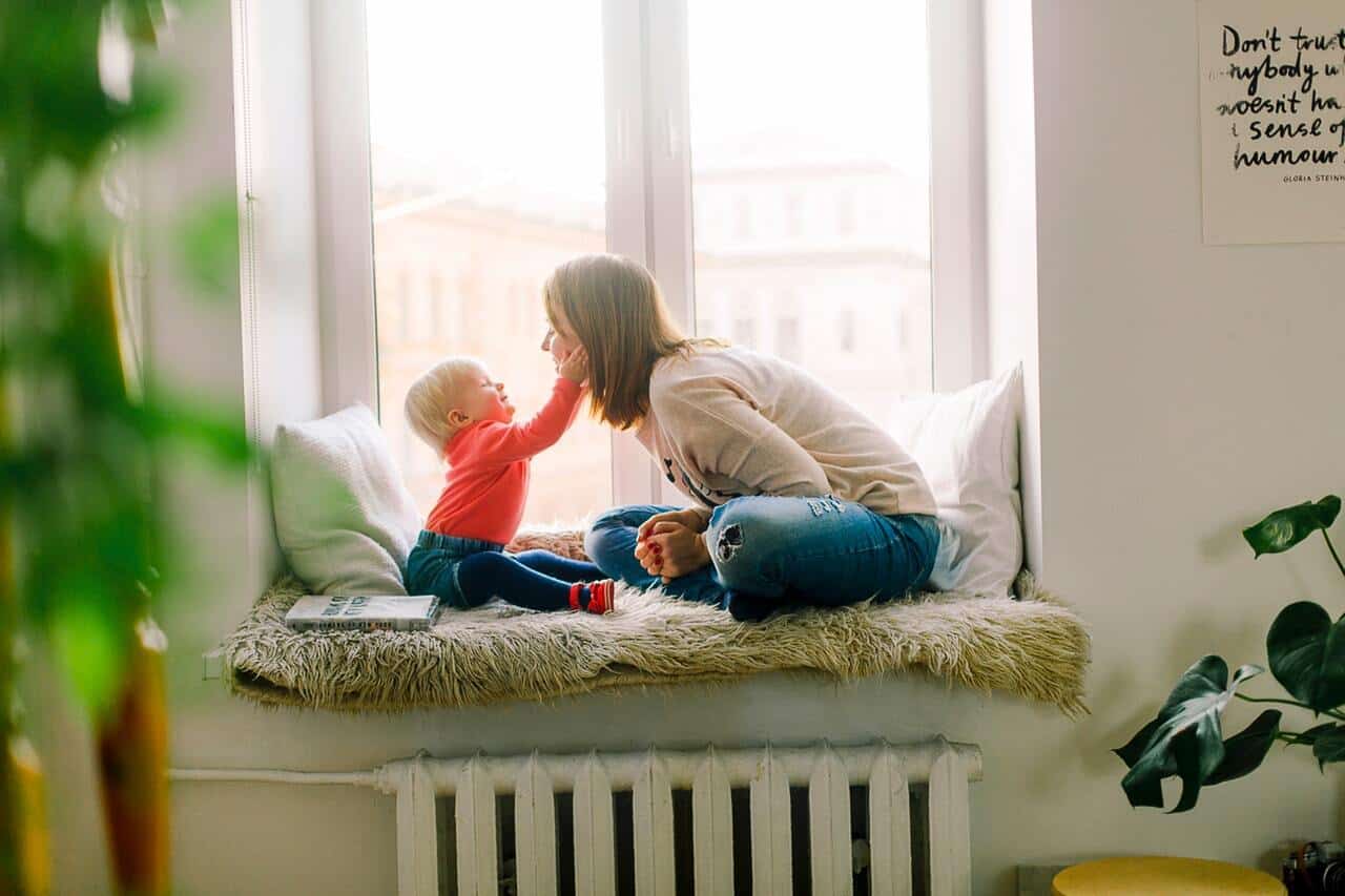 A baby holding its mother's cheeks in their hands upon a windowsill