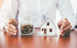 Saving money for a house, person in white shirt sitting at wood table. Their hands are on either side of a jar of saved coins and a miniature model of a house.