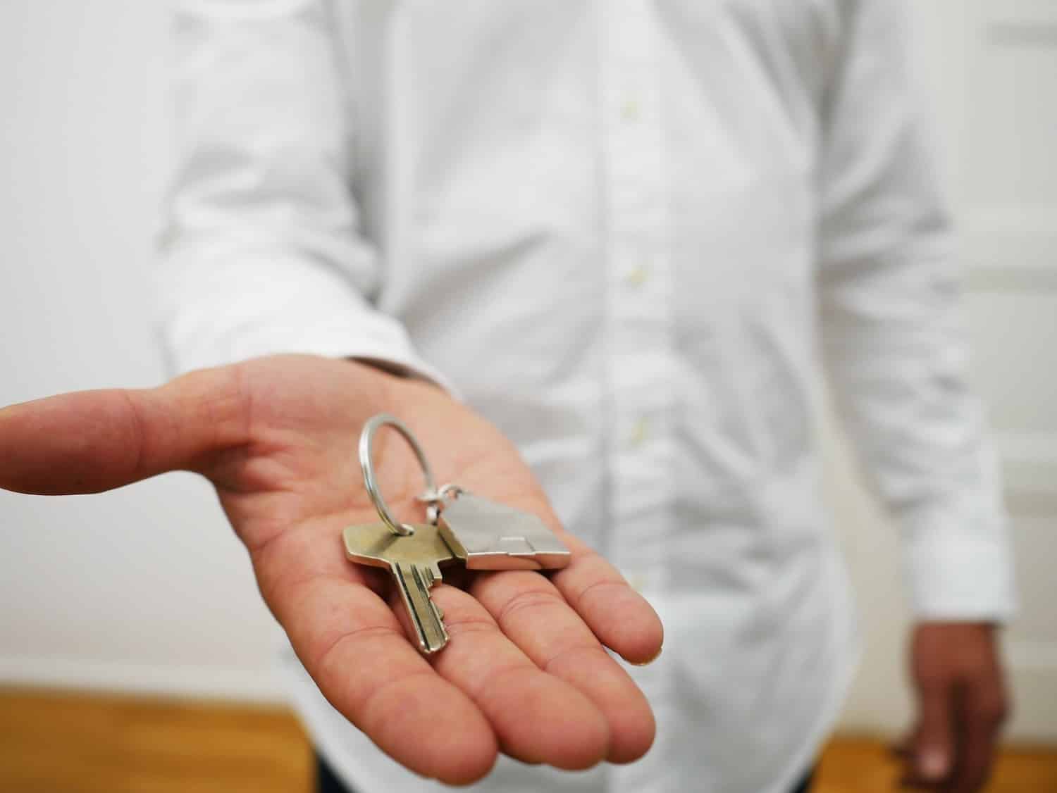 Person in white button-up extending hand toward camera, a house key with a house-shaped keychain in their palm