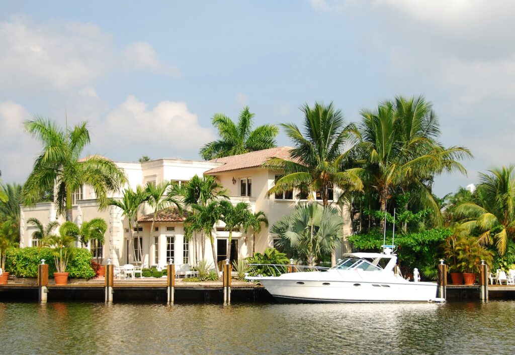 A waterfront mansion with decorative palm trees and a small yacht by the dock.