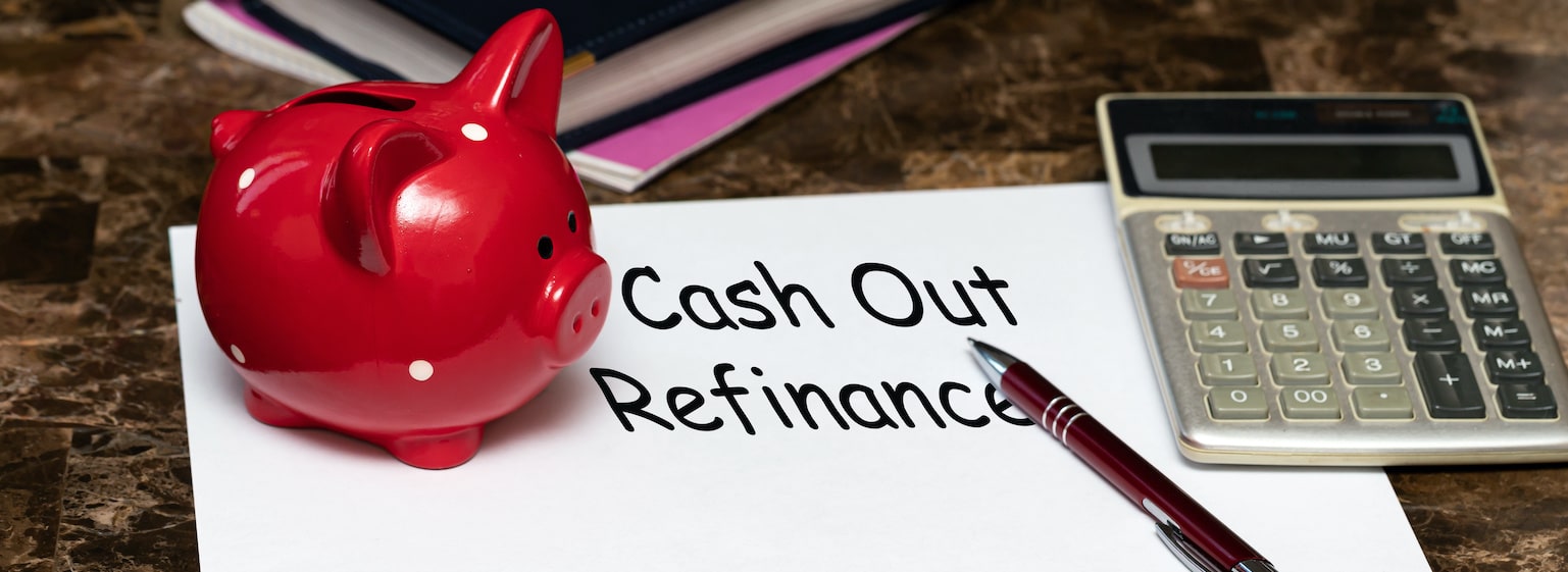 Paper with the words cash out refinancing on a table next to a red piggy Bank. Cash-Out Refinancing: Covering the Pros and Cons