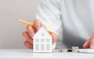 A person holding a pencil near a miniature house model and calculating savings with stacked coins, representing strategies to lower mortgage payments.