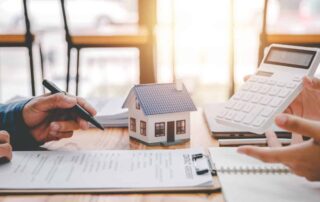 Two individuals discussing home loan refinancing with a miniature house model, documents, and a calculator, focusing on VA loan refinancing options.